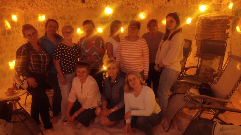 Grupa jedenastu uśmiechniętych kobiet pozuje wspólnie w jaskini solnej. Pomieszczenie jest wypełnione białym puchem solnym na podłodze i oświetlone ciepłym, pomarańczowym światłem lamp solnych na ścianach