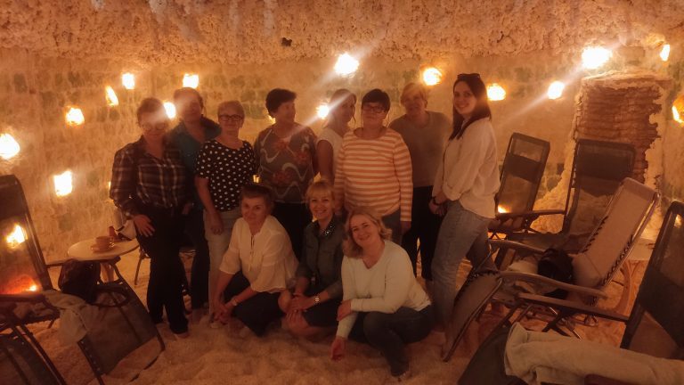 Grupa jedenastu uśmiechniętych kobiet pozuje wspólnie w jaskini solnej. Pomieszczenie jest wypełnione białym puchem solnym na podłodze i oświetlone ciepłym, pomarańczowym światłem lamp solnych na ścianach