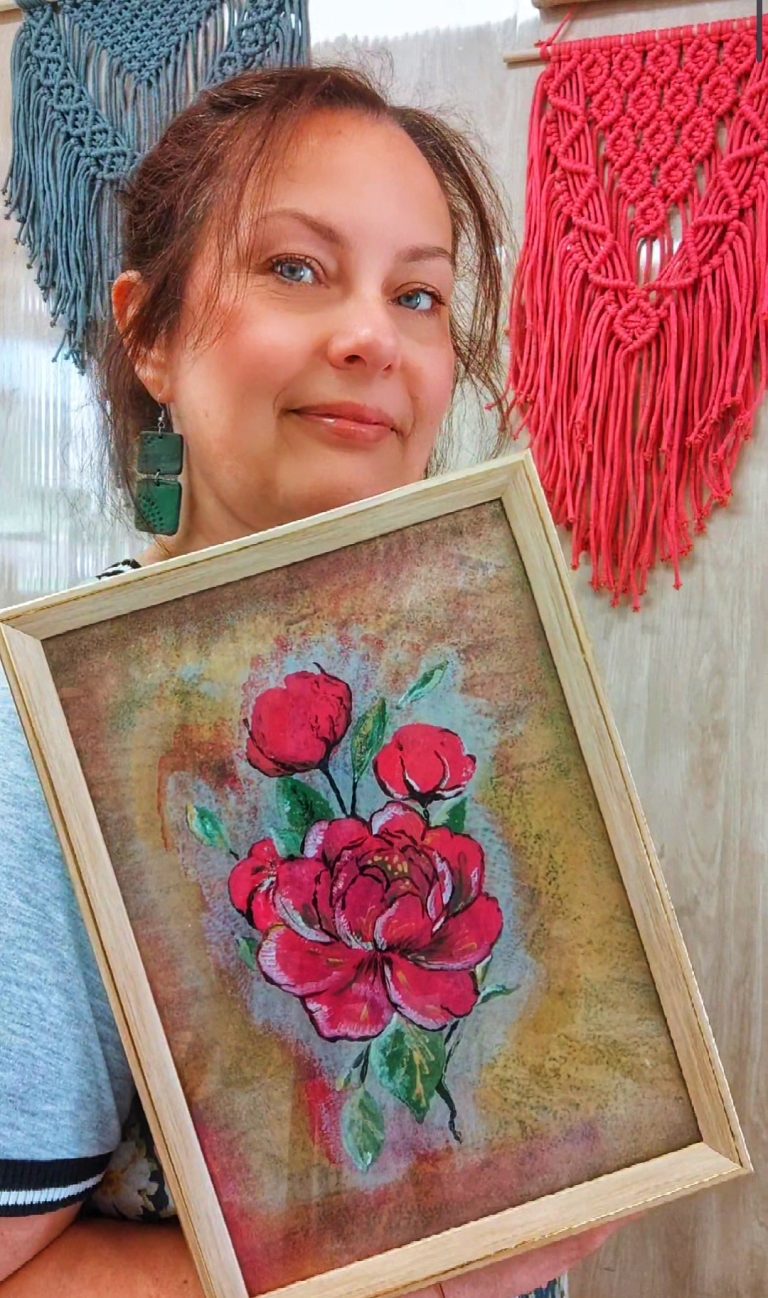 Portret Moniki Wojdyło trzymającej oprawiony w drewnianą ramkę obraz przedstawiający czerwone kwiaty piwonii. W tle widoczne są ozdobne makramy ścienne w kolorze szarym i różowym