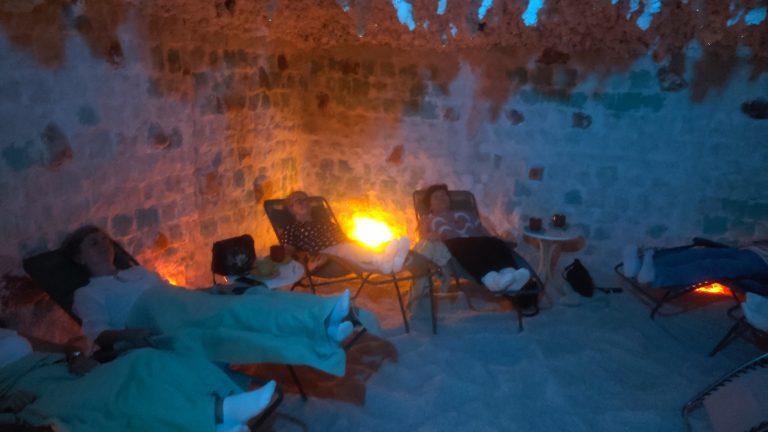 Cztery kobiety odpoczywają na leżakach w jaskini solnej. Pomieszczenie ma białe ściany z bloków soli, podłogę pokrytą solą oraz nastrojowe, punktowe oświetlenie w kolorze pomarańczowym i niebieskim. Uczestniczki są przykryte jasnymi kocami