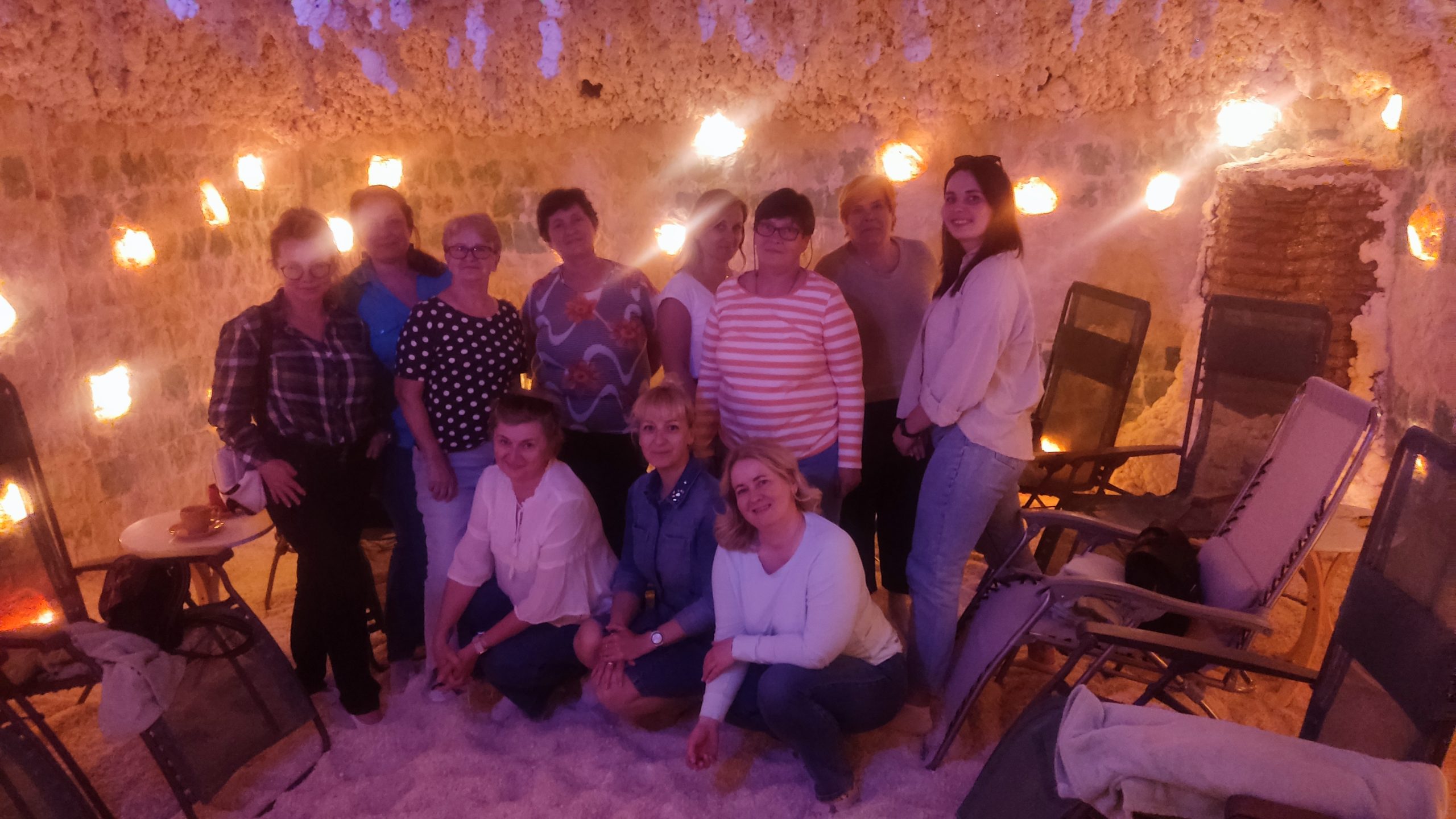 Grupa jedenastu uśmiechniętych kobiet pozuje wspólnie w jaskini solnej. Pomieszczenie jest wypełnione białym puchem solnym na podłodze i oświetlone ciepłym, pomarańczowym światłem lamp solnych na ścianach