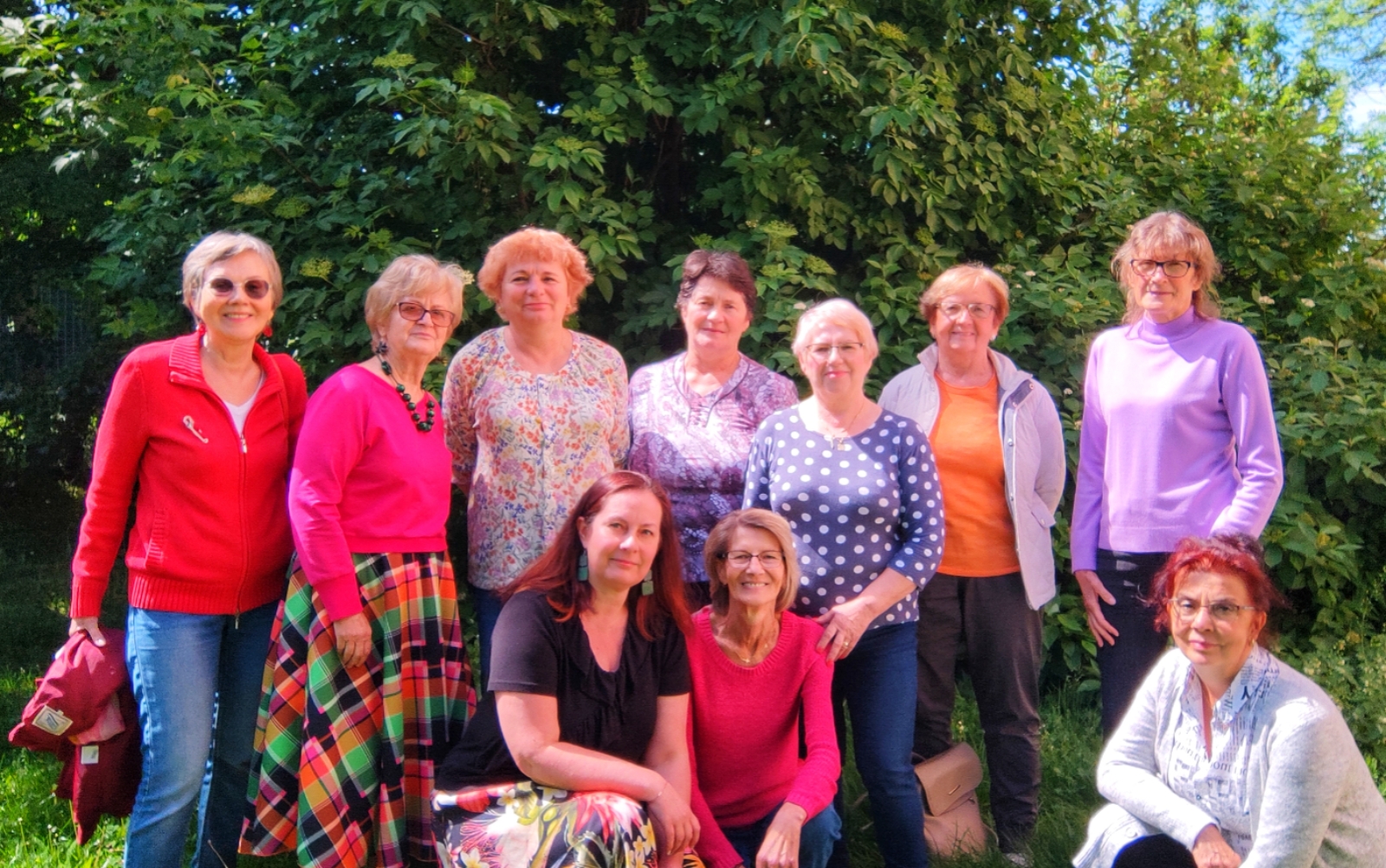 Grupa jedenastu uśmiechniętych kobiet pozuje do wspólnego zdjęcia w słonecznym ogrodzie na tle gęstych, zielonych krzewów. Uczestniczki są ubrane w kolorowe, wiosenne stroje – dominują odcienie czerwieni, różu, fioletu i wzory kwiatowe. Monika Wojdyło klęczy w pierwszym rzędzie po lewej stronie.