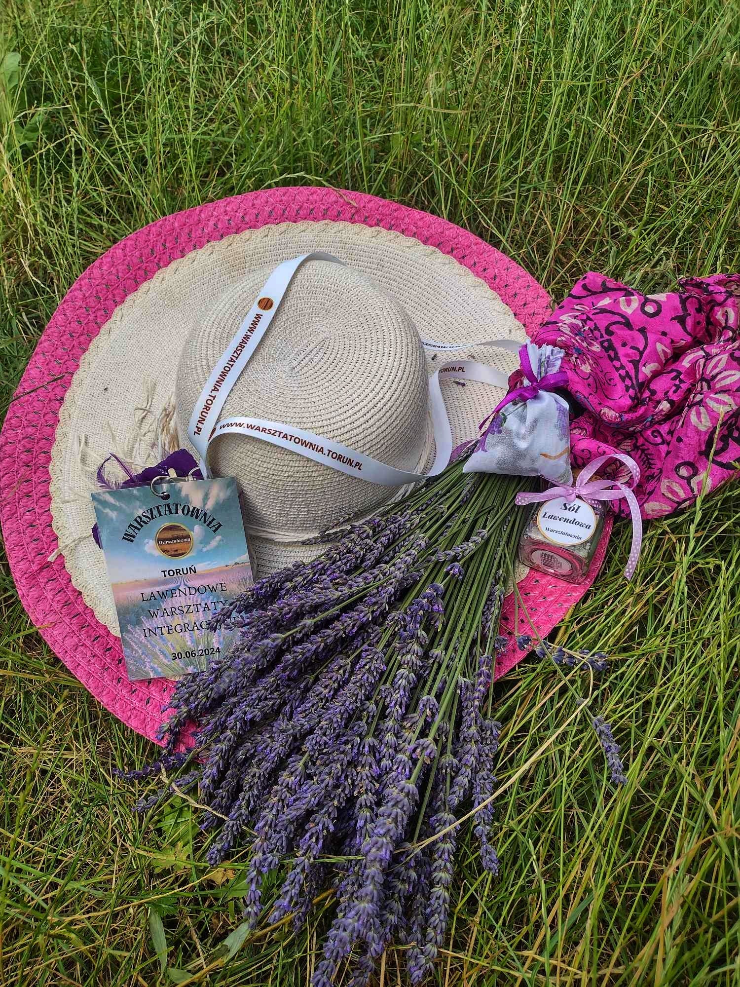Słomkowy kapelusz z szerokim różowym rondem leży na trawie obok dużego bukietu świeżej lawendy, słoiczka soli lawendowej Warsztatowni oraz identyfikatora z napisem „Lawendowe Warsztaty Integracyjne”