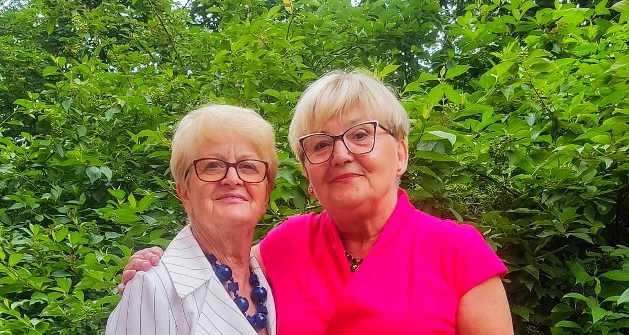 Dwie uśmiechnięte kobiety w wieku dojrzałym, ubrane w eleganckie stroje, pozują na tle gęstych, zielonych krzewów. Jedna ma na sobie białą marynarkę w prążki i efektowny, duży granatowy naszyjnik, a druga intensywnie różową bluzkę i subtelną biżuterię