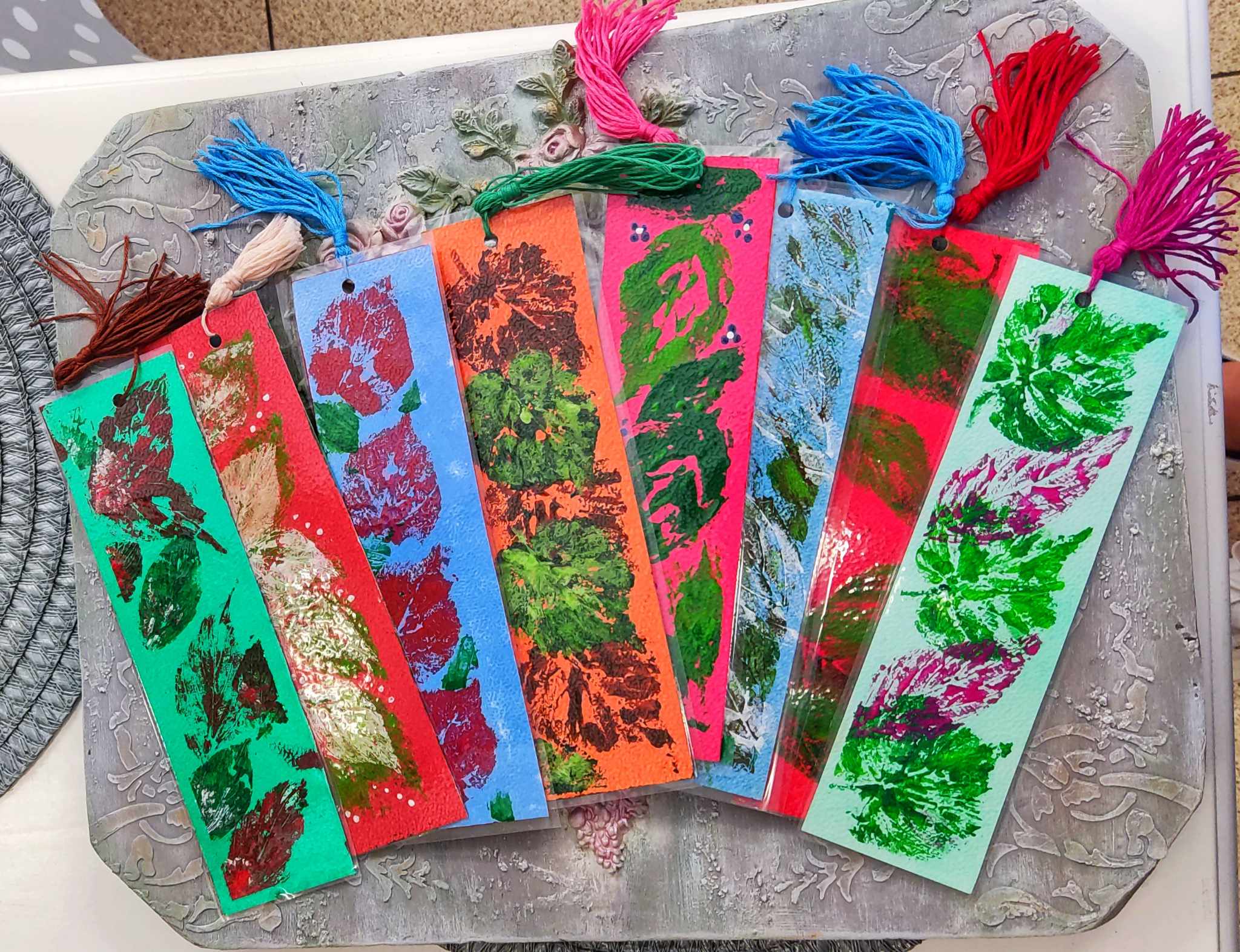Siedem ręcznie wykonanych, kolorowych zakładek do książek z chwostami, ozdobionych motywami roślinnymi wykonanymi techniką odciskania liści. Zakładki mają intensywne barwy: czerwoną, niebieską, pomarańczową, różową i zieloną