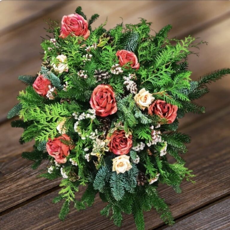 Okrągła, gęsta wiązanka nagrobna na ciemnym, drewnianym stole. Kompozycja wykonana z zielonych gałązek świerku i tui, ozdobiona licznymi czerwonymi różami, drobnymi beżowymi kwiatuszkami oraz naturalnymi szyszkami