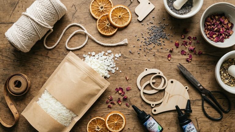 Artystyczne ujęcie typu flatlay na drewnianym stole, przedstawiające naturalne materiały do rękodzieła: otwartą torebkę z płatkami wosku sojowego, suszone plastry pomarańczy, suszoną lawendę i płatki róż w miseczkach, bawełniany sznurek do makramy, butelki z żywicą UV, drewniane elementy oraz nożyczki w stylu vintage