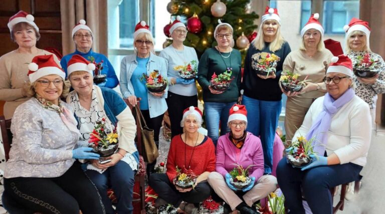 Duża grupa seniorek w mikołajkowych czapkach prezentuje gotowe dekoracje bożonarodzeniowe podczas warsztatów Warsztatowni