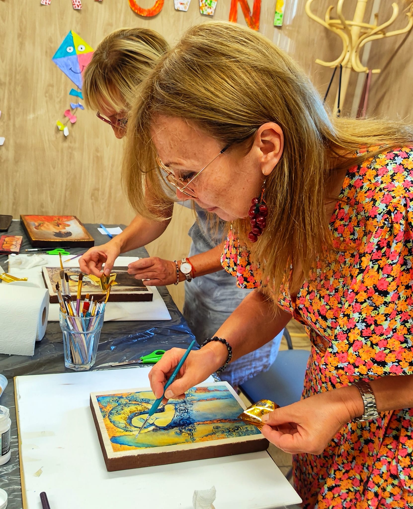 Uczestniczka warsztatów w Warsztatowni nakłada płatki złota na obraz przy użyciu pędzla; na stole widać profesjonalne akcesoria malarskie