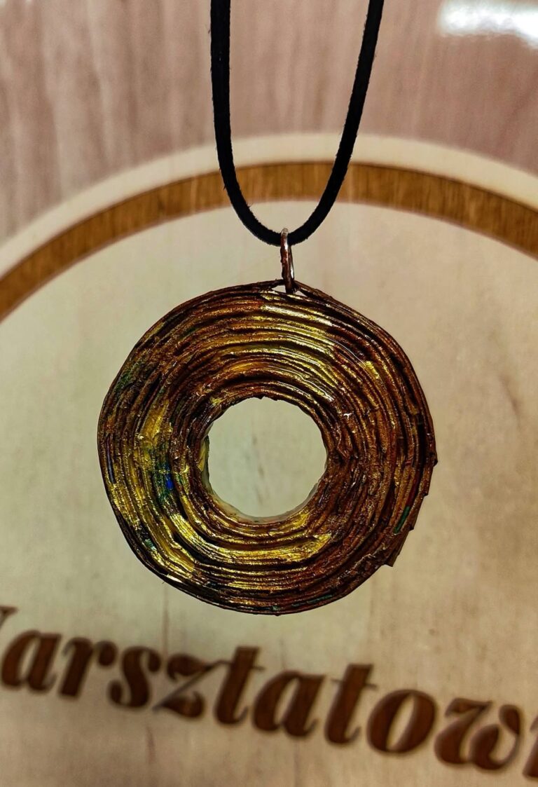 Okrągły wisior z otworem w środku, wykonany z ciasno zwiniętej wikliny papierowej o wyraźnej, warstwowej strukturze. Biżuteria jest pomalowana na metaliczny, złoty kolor z widocznymi przetarciami w odcieniach miedzi, zieleni i błękitu. Wisior wisi na cienkim, czarnym rzemyku na tle jasnego drewna z fragmentem napisu "Warsztatownia"
