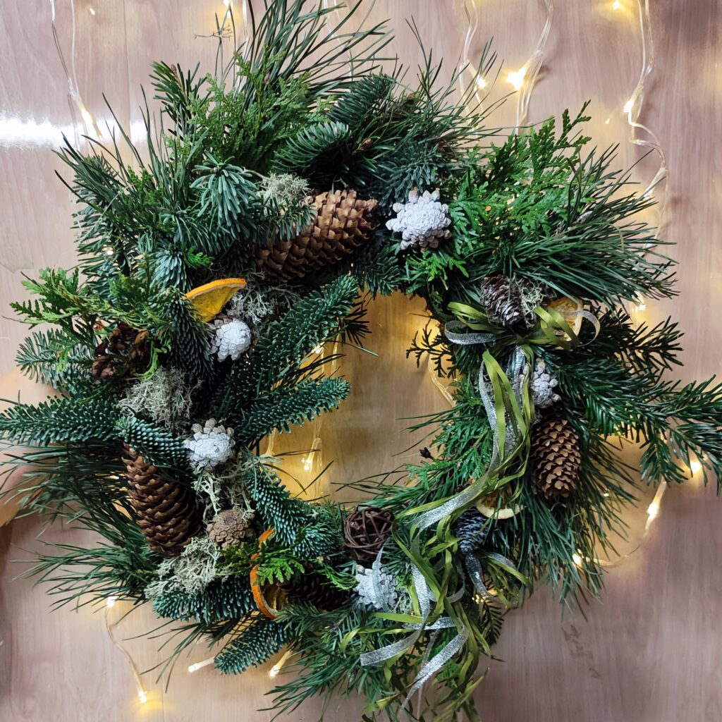 Bożonarodzeniowy wieniec dekoracyjny z gałązek choinkowych, szyszek, pomarańczy, białych kulek i zielonej wstążki, podświetlony lampkami na drewnianym tle