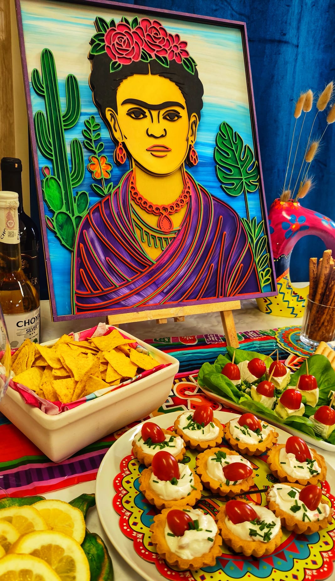 Relief 3D z Fride Kahlo na tle niebieskim stojący obok stołu z przekąskami: nachosami, pomidorami cherry z serem, tortille, cytrynami, butelką tequili i dekoracjami meksykańskimi