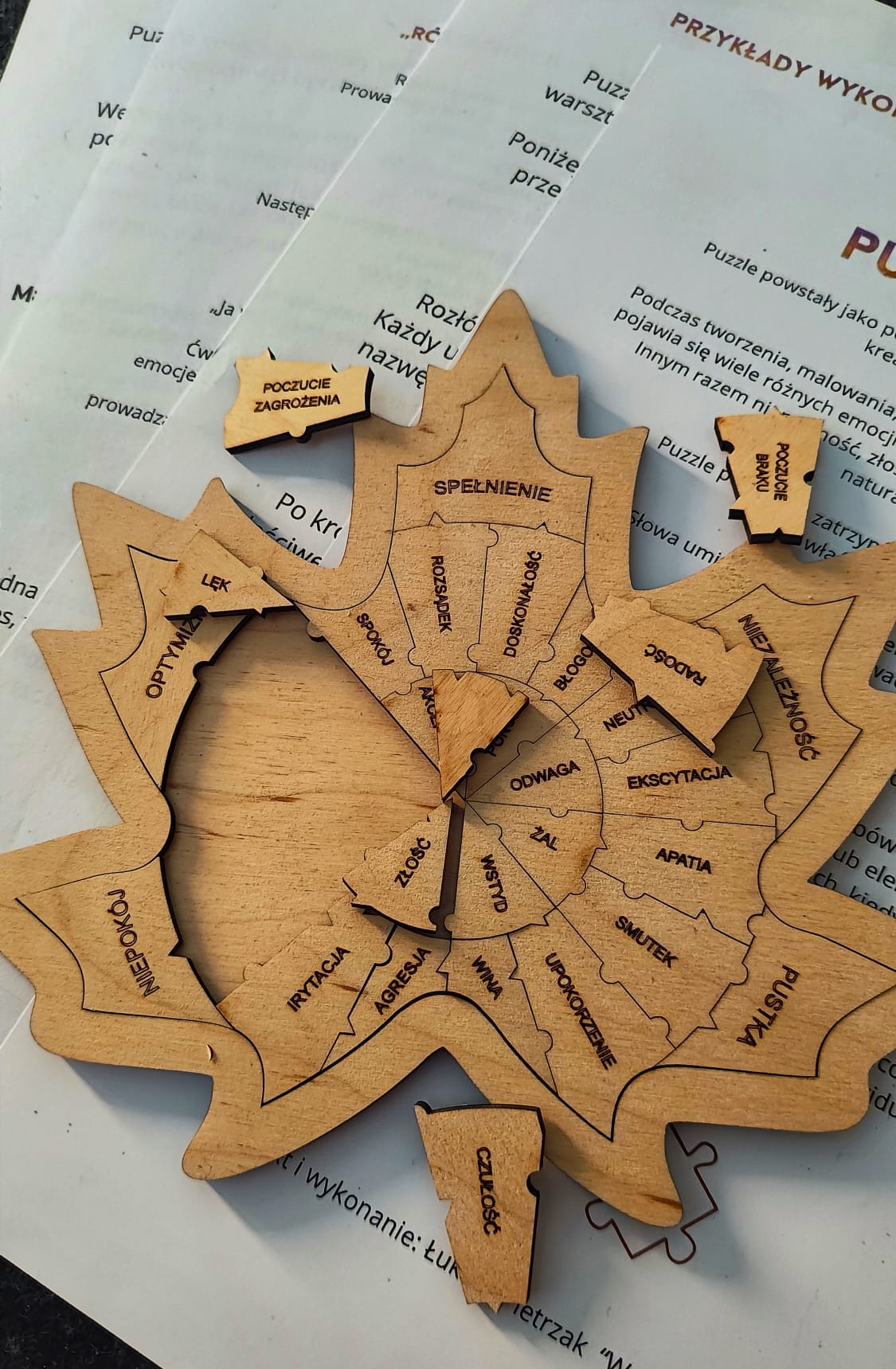 Drewniane „puzzle emocji” w kształcie liścia, ułożone na wydrukowanych kartach z instrukcją i opisem pracy z emocjami na szarym tle