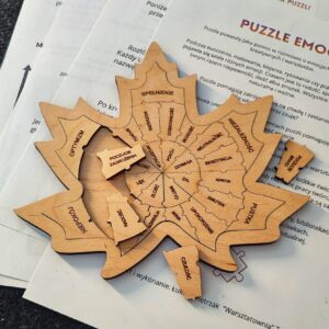 Drewniane „puzzle emocji” w kształcie liścia, ułożone na wydrukowanych kartach z instrukcją i opisem pracy z emocjami na szarym tle