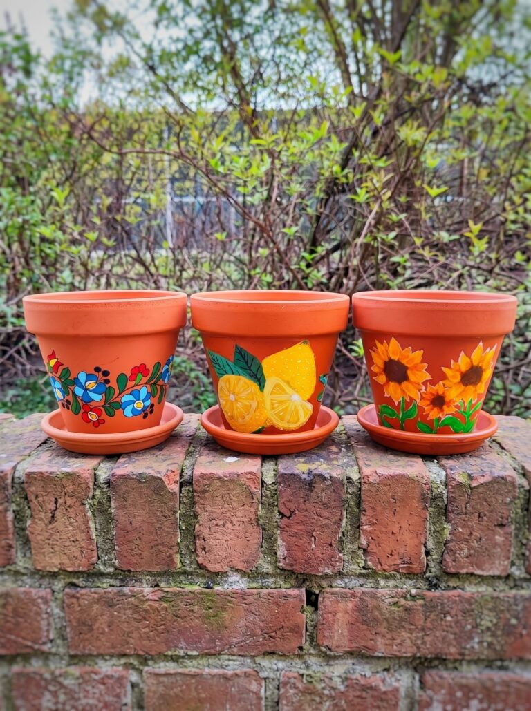 Trzy pomalowane doniczki z terakoty stojące na ceglanej ścianie na tle ogrodu. Każda ma inny ręcznie malowany wzór: kwiaty w stylu ludowym, cytryny oraz słoneczniki