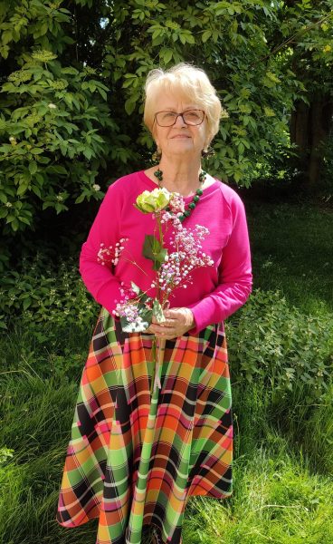 Portret uśmiechniętej seniorki w okularach i różowej bluzce, trzymającej bukiet kolorowych kwiatów (gipsówki i róży). Kobieta stoi w słonecznym ogrodzie na tle zielonych krzewów, mając na sobie kolorową spódnicę w kratę i zielone korale
