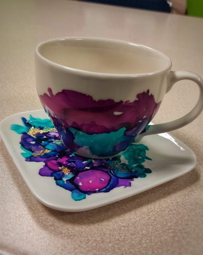 Biała ceramiczna filiżanka i kwadratowy spodek udekorowane abstrakcyjnym wzorem wykonanym tuszami alkoholowymi w odcieniach intensywnego błękitu, turkusu i fioletu. Wzór przypomina płynne, przenikające się plamy barwne, które są dodatkowo ozdobione nieregularnymi akcentami złotego szlagmetalu. Zestaw stoi na jasnej, drewnianej półce na tle drewnianej ściany
