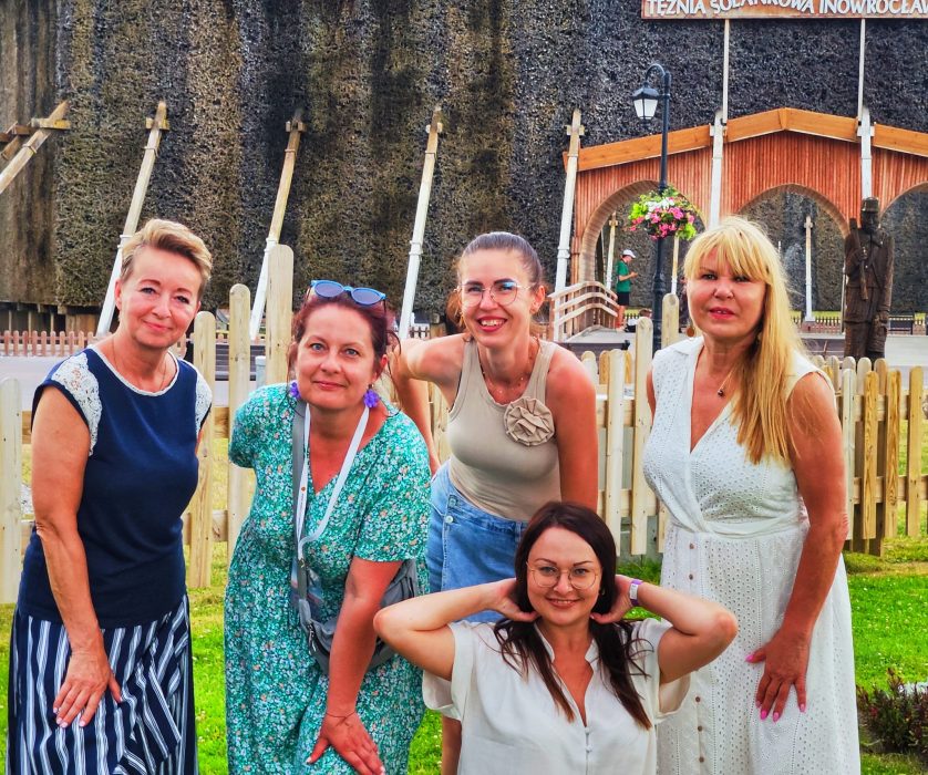 Pięć uśmiechniętych kobiet pozuje na tle drewnianej tężni solankowej w Inowrocławiu. Monika Wojdyło w zielonej sukience stoi obok uczestniczek w letnich stylizacjach
