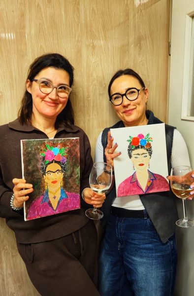 Dwie uśmiechnięte kobiety w okularach pozujące z ukończonymi obrazami Fridy Kahlo na płótnach, trzymając kieliszki wina, na tle drewnianej ściany w Warsztatowni