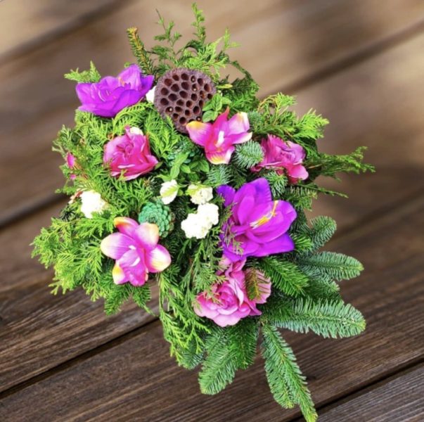 Pionowa wiązanka nagrobna na drewnianym stole, wykonana z zielonych gałązek świerku i tui. Kompozycja ozdobiona jest kwiatami w odcieniach intensywnego różu i fioletu oraz naturalnym suszem lotosu umieszczonym w centralnej części