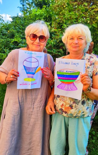 Dwie uśmiechnięte kobiety w okularach przeciwsłonecznych stoją na tle zieleni, trzymając kartki z kolorowymi rysunkami filiżanek opisanych jako Miłość, Radość i Zadowolenie