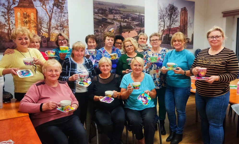 Duża grupa uśmiechniętych kobiet prezentuje własnoręcznie pomalowane, kolorowe filiżanki i spodeczki podczas warsztatów rzemieślniczych