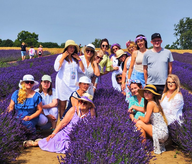 Duża grupa uśmiechniętych osób w różnym wieku, ubranych w kolorowe letnie stroje i kapelusze, pozuje wspólnie pośród rzędów kwitnącej lawendy pod błękitnym niebem