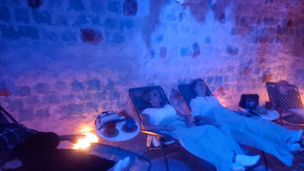 Cztery kobiety odpoczywają na leżakach w jaskini solnej. Pomieszczenie ma białe ściany z bloków soli, podłogę pokrytą solą oraz nastrojowe, punktowe oświetlenie w kolorze pomarańczowym i niebieskim. Uczestniczki są przykryte jasnymi kocami