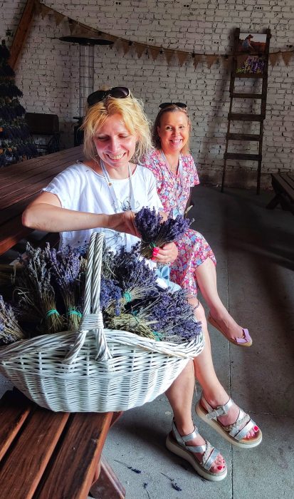 Duży, biały wiklinowy kosz wypełniony po brzegi bukietami suszonej, fioletowej lawendy. W tle dwie uśmiechnięte kobiety siedzące na ławce w rustykalnym wnętrzu