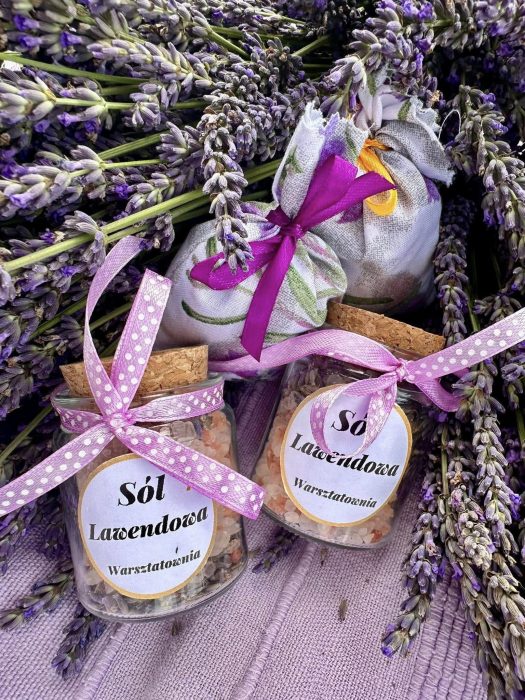 Kompozycja na fioletowym tle: dwa szklane słoiczki z solą lawendową Warsztatowni ozdobione różowymi wstążkami, dwa płócienne woreczki zapachowe z fioletowymi kokardami oraz bukiet świeżo ściętej lawendy