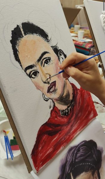 Makro zdjęcie ręki malującej twarz Fridy Kahlo na płótnie podczas warsztatów, z widocznym niebieskim pędzlem, konturami oczu i ust, czerwonym strojem oraz innymi obrazami w tle