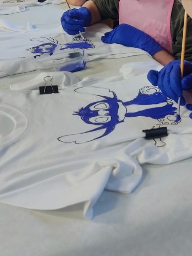 Biała koszulka dziecięca zawieszona na czarnym wieszaku, ozdobiona ręcznie malowaną postacią Stitcha w intensywnych kolorach niebieskim i różowym. Postać ma duże oczy, podniesione uszy i siedzi centralnie na froncie koszulki. W tle widać kolorowe dekoracje urodzinowe, co sugeruje kreatywne zajęcia podczas przyjęcia