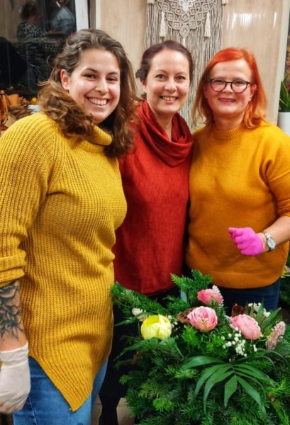 Trzy uśmiechnięte kobiety w kolorowych swetrach (żółtym, czerwonym i musztardowym) stoją nad zieloną wiązanką nagrobną. Środkowa postać, Monika Wojdyło, uśmiecha się do obiektywu, wspierając uczestniczki w pracy twórczej