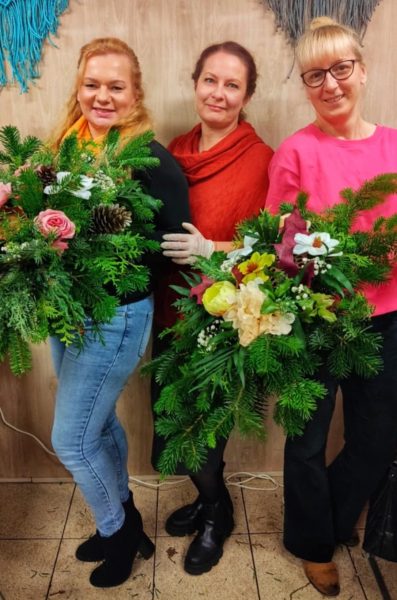 Trzy uśmiechnięte kobiety stoją obok siebie w pracowni, trzymając duże, własnoręcznie wykonane wiązanki nagrobne z żywego igliwia i kolorowych kwiatów. Środkowa postać to Monika Wojdyło w czerwonym stroju