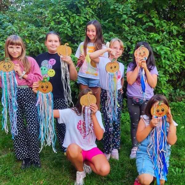 Grupa siedmiu dziewczynek pozuje w ogrodzie, trzymając własnoręcznie wykonane dekoracje – uśmiechnięte dynie z kolorowymi sznurkami do makramy. Niektóre dzieci żartobliwie zasłaniają twarze swoimi pracami