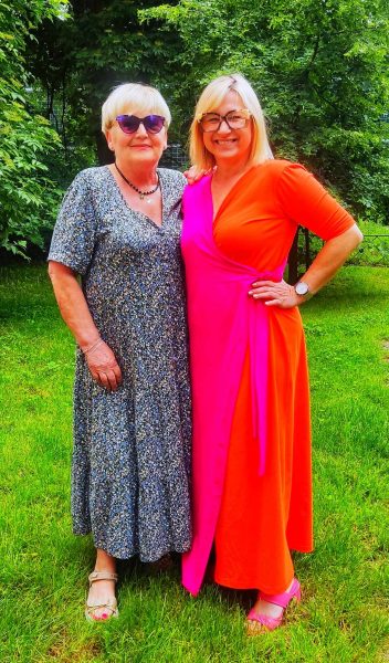 Dwie uśmiechnięte kobiety stoją obok siebie w ogrodzie. Kobieta po lewej ma okulary przeciwsłoneczne i długą sukienkę w drobne kwiaty. Kobieta po prawej, Monika Wojdyło, ubrana jest w długą, dwukolorową sukienkę (różowo-pomarańczową) i uśmiecha się promiennie