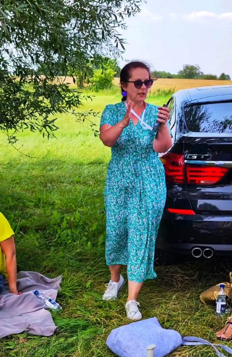 Monika Wojdyło w zielonej sukience w kwiaty stoi przy czarnym samochodzie na polanie i gestykulując, opowiada grupie o właściwościach soli inowrocławskiej