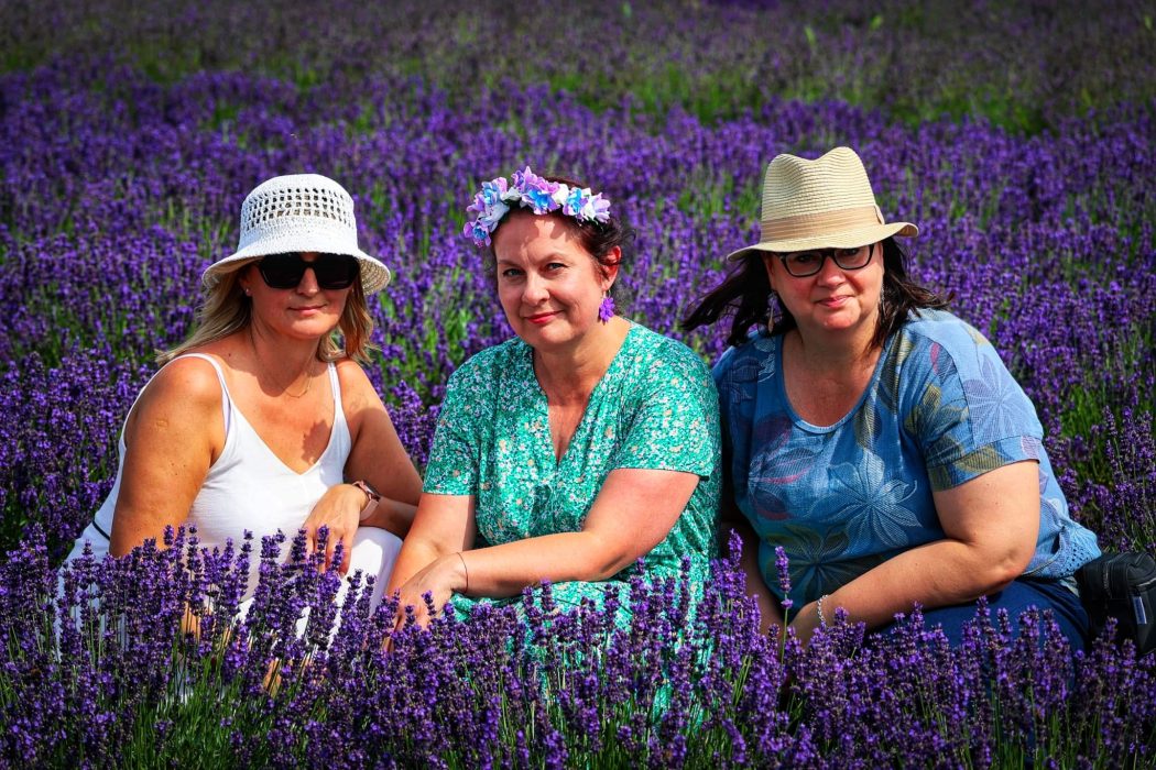 Trzy uśmiechnięte kobiety siedzą pośród gęstych rzędów kwitnącej lawendy. W środku Monika Wojdyło w zielonej sukience w drobne kwiaty i fioletowym wianku na głowie, otoczona dwiema uczestniczkami w kapeluszach