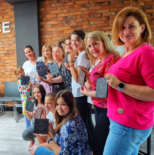 Duża grupa uśmiechniętych kobiet i dzieci w różnym wieku, pozująca z gotowymi pracami malowanymi metodą kropkową. Monika Wojdyło stoi na końcu grupy po lewej stronie, a uczestniczki trzymają przed sobą czarne, kwadratowe obrazy z kolorowymi mandalami. Tło stanowi nowoczesna ściana z czerwonej cegły