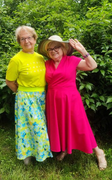 Dwie uśmiechnięte kobiety w wieku dojrzałym pozują w ogrodzie. Kobieta po lewej ma na sobie neonowo-żółty t-shirt z napisem i błękitną spódnicę w kwiaty. Kobieta po prawej, w słomkowym kapeluszu i okularach, ubrana jest w intensywnie różową sukienkę i radośnie macha do obiektywu