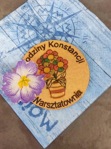 Dekoracyjny magnes lub pamiątkowa tabliczka z niebieskim, przecieranym tłem i okrągłą, żółtą etykietą pośrodku. Na etykiecie widnieje napis „Urodziny Konstancji” oraz „Warsztatownia”, a obok znajduje się grafika doniczki z kolorowymi kwiatami. W lewym dolnym rogu naklejono dużą, prawdziwą lub sztuczną fioletowo-białą kwiatową ozdobę