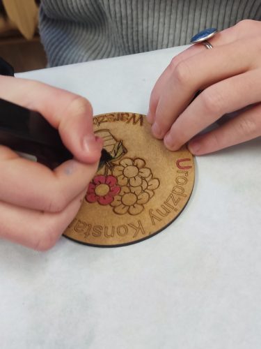Dekoracyjny magnes lub pamiątkowa tabliczka z niebieskim, przecieranym tłem i okrągłą, żółtą etykietą pośrodku. Na etykiecie widnieje napis „Urodziny Konstancji” oraz „Warsztatownia”, a obok znajduje się grafika doniczki z kolorowymi kwiatami. W lewym dolnym rogu naklejono dużą, prawdziwą lub sztuczną fioletowo-białą kwiatową ozdobę