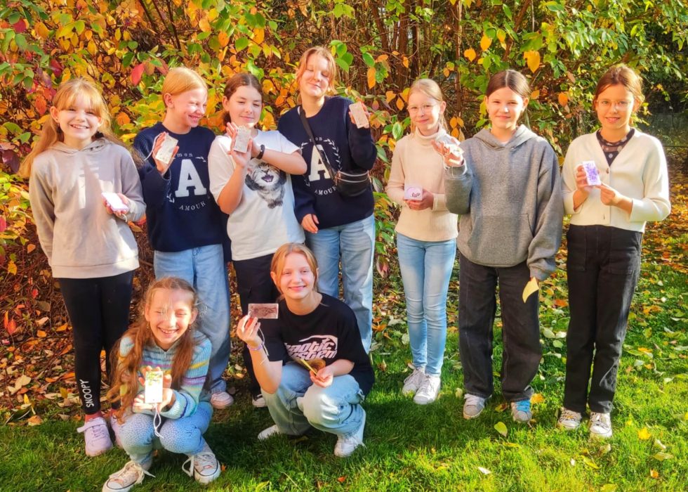 Grupa uśmiechniętych dziewczynek pozuje w słonecznym ogrodzie, trzymając w dłoniach własnoręcznie wykonane pamiątki z warsztatów rękodzielniczych. W tle jesienne liście i krzewy