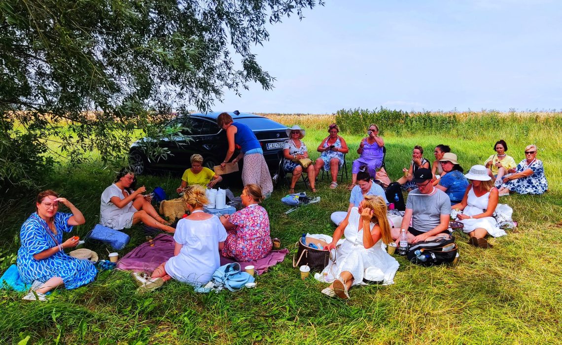 Duża grupa uczestników wycieczki odpoczywa na kocach w cieniu rozłożystego drzewa na zielonej polanie. W tle widoczny czarny samochód oraz złote pole zboża pod błękitnym niebem