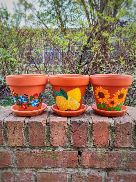 Trzy pomalowane doniczki z terakoty stojące na ceglanej ścianie na tle ogrodu. Każda ma inny ręcznie malowany wzór: kwiaty w stylu ludowym, cytryny oraz słoneczniki