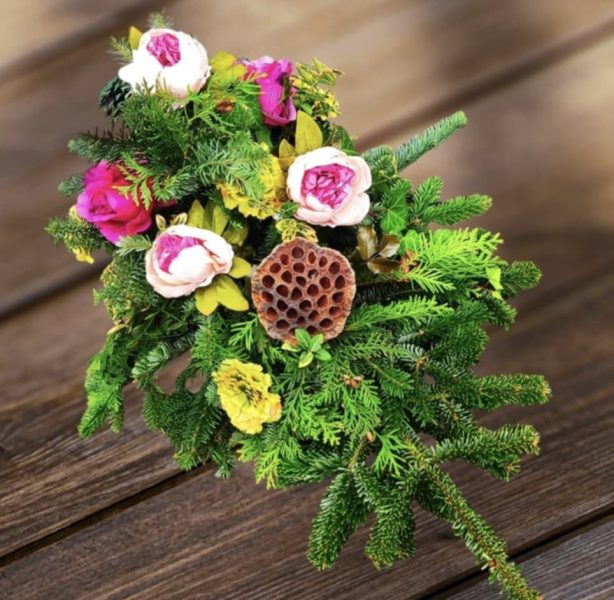 Profesjonalna wiązanka nagrobna leżąca na drewnianym stole, wykonana z gęstych gałązek zielonego świerku, ozdobiona jasnoróżowymi różami, żółtymi goździkami oraz charakterystycznym, brązowym suszem lotosu
