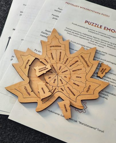 Drewniane „puzzle emocji” w kształcie liścia, ułożone na wydrukowanych kartach z instrukcją i opisem pracy z emocjami na szarym tle