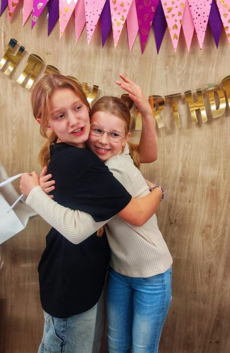 Dwie uśmiechnięte dziewczynki tulące się do siebie na tle dekoracji urodzinowych. W tle widoczna różowo-fioletowa girlanda z chorągiewek oraz złoty napis „HAPPY BIRTHDAY” na drewnianej ścianie