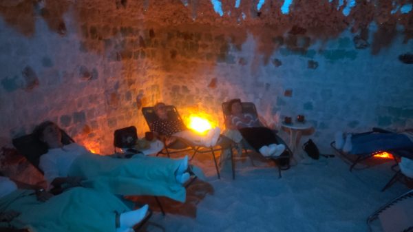 Cztery kobiety odpoczywają na leżakach w jaskini solnej. Pomieszczenie ma białe ściany z bloków soli, podłogę pokrytą solą oraz nastrojowe, punktowe oświetlenie w kolorze pomarańczowym i niebieskim. Uczestniczki są przykryte jasnymi kocami