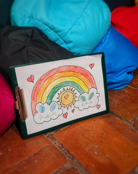 Kolorowy rysunek tęczy z uśmiechniętym słońcem w środku i dwiema chmurkami (jedna radosna, druga smutna), przypięty do zielonej podkładki z klipsem. Wokół tęczy narysowane są małe czerwone serduszka. Podkładka opiera się o kolorowe pufy sako na ceglanej podłodze
